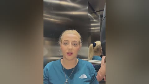 Video of a blonde, light-skinned woman in a blue scrub top, smiling, in a modern, stainless steel hospital hallway with blurred background.