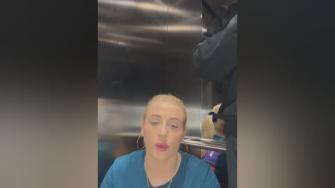 Video of a blonde woman with hoop earrings in a teal scrub top, sitting in a dimly lit, sterile medical room with metallic walls and equipment.