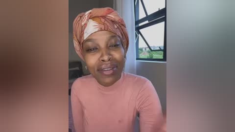 Media: Video of a young woman with a light brown complexion, wearing a pink long-sleeve shirt and a patterned headscarf. She has a neutral expression and is indoors with a window behind her.