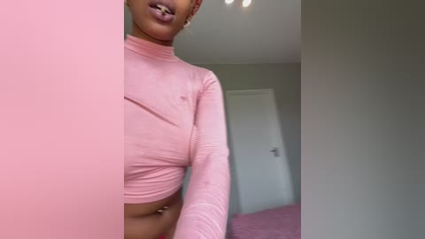 Media: A video of a young, dark-skinned woman with short hair wearing a tight, pink crop top and pink lipstick, standing in a minimalistic, softly lit room with a pink carpet.