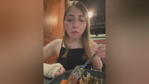 Media: A video of a young woman with light skin and long brown hair, wearing a black tank top, eating from a black bowl in a dimly lit restaurant with wooden walls.