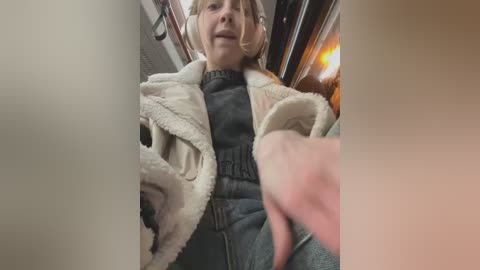 Media: Video of a blonde woman in a beige fur coat, black sweater, and jeans, gesturing with her right hand in a subway car, lit by warm interior lighting.