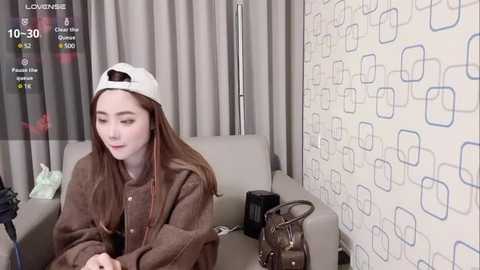 Media: Video of a young Asian woman with long, straight brown hair, wearing a white baseball cap and brown jacket, sitting on a gray couch in a modern room with patterned wallpaper.