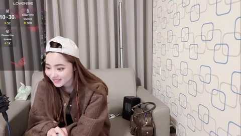 Media: Video of a young woman with light skin and straight brown hair, wearing a white cap and brown jacket, smiling while seated on a gray couch in a modern room with geometric-patterned wallpaper.