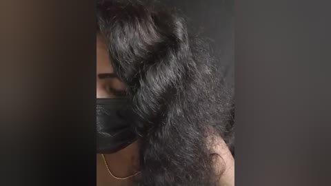 Media: A close-up video of a person with dark skin and long, voluminous, wavy black hair partially covering their face. They are wearing a black mask, which obscures their nose and mouth, and a gold chain necklace. The background is dark and blurry, focusing attention on the hair and mask.