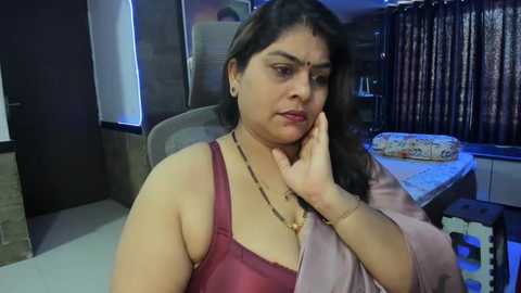 Media: Video of a South Asian woman with medium skin tone, long dark hair, wearing a pink blouse and gold necklace, sitting in a modern, dimly lit living room with a sofa, curtains, and a decorative pillow.