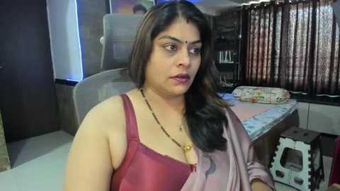 A video shows a woman with medium skin tone and long, dark hair in a maroon top, sitting at a desk with a patterned tablecloth and books in a dimly lit room.