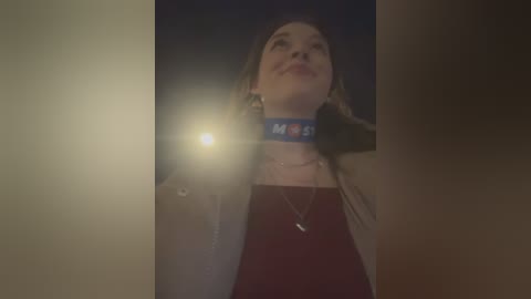 Media: Video of a smiling young woman with blonde hair, wearing a red top and beige jacket, standing outside at night with a flashlight.