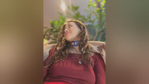 Video of a woman with wavy brown hair, wearing a red sweater and a blue collar, sitting in a cozy, dimly lit room with green plants in the background.