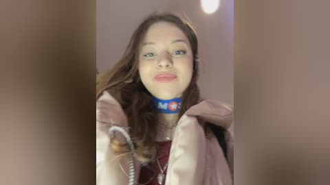 Video of a young woman with light skin and long brown hair, wearing a blue choker with red and white letters, a pink jacket, and a red shirt.