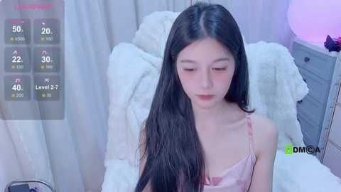 Media: Video of a young Asian woman with long black hair, wearing a light pink camisole, sitting on a white bed with fluffy pillows. The background features a bedside table with a lamp and digital temperature display.