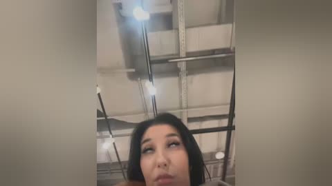 Media: Video of a young Asian woman with dark hair, wearing makeup, looking upward in a dimly lit, industrial room with exposed beams and electrical wires.