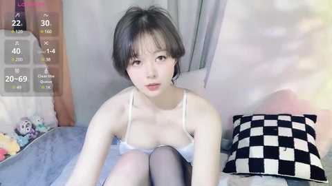 Media: A video of an Asian woman with short black hair, fair skin, and a slender physique, wearing a white bra and black stockings, sitting on a bed with a checkered pillow and plush toys in the background.