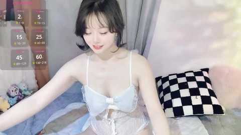 Media: Video of an East Asian woman with fair skin, short black hair, wearing a white lace lingerie set, sitting on a bed with a black-and-white checkered pillow, surrounded by stuffed toys.