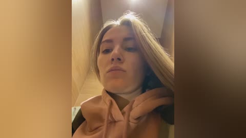 A video of a young Caucasian woman with light skin and straight, shoulder-length blonde hair. She's wearing a light pink hoodie and has a contemplative expression. The background is a dimly lit hallway with beige walls.