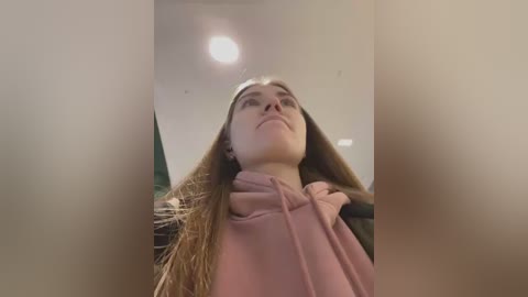 Video of a young Caucasian woman with long brown hair, wearing a pink hoodie, looking up with a contemplative expression, against a blurred indoor background.