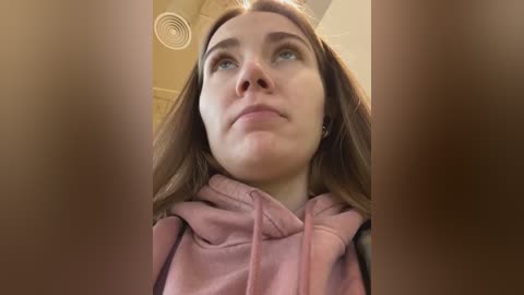 Media: A video of a young woman with fair skin and long brown hair, wearing a pink hoodie, looking up with a thoughtful expression in a dimly lit room with a ceiling vent.