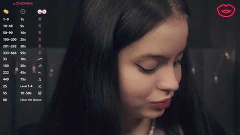 Media: Video of a young woman with long, straight black hair, light skin, and minimal makeup, wearing a black top, in a dimly lit room. The background shows a dark wall and a digital interface with various icons.