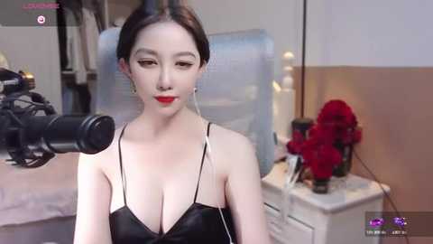 Media: Video of a fair-skinned East Asian woman with dark hair, wearing a black, low-cut top, speaking into a microphone in a bedroom with a bed, a white dresser, and a vase of red roses.