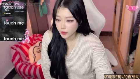 Media: A video of an East Asian woman with long black hair, wearing a beige knitted sweater, sitting on a white chair in a pink room. A striped pillow with a cartoon character is visible behind her.