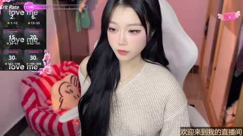 Media: Video of an East Asian woman with long black hair in a beige sweater, sitting on a white gaming chair. In the background, a pink wall, a stuffed toy, and a rating screen displaying \"Love Me.\