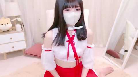 Media: Video of an Asian woman with long black hair, wearing a white mask, white off-shoulder top, red bow, and red skirt, sitting on a plush rug in a minimalist room with a white chair, dresser, and mirror.