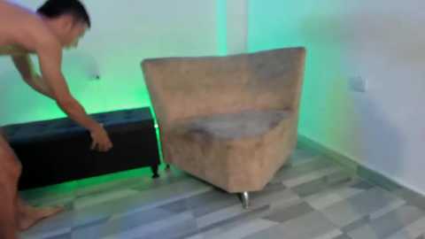 Video of a nude man with short dark hair bending over to lift a large, beige, angular armchair with light wood legs. Background features a white wall, a black ottoman, and a green neon light.