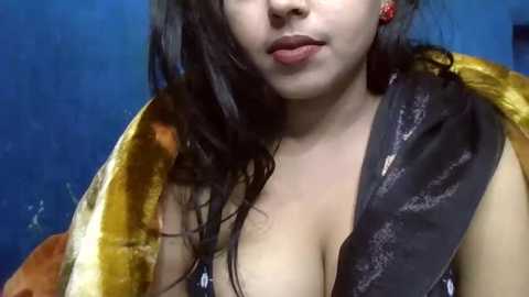 Media: Video of a light-skinned woman with long, wavy black hair, wearing a yellow blouse with a plunging neckline, revealing cleavage, and a black shawl. Background is a textured blue wall.