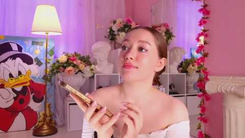 Media: Video of a young woman with fair skin and brown hair in an off-the-shoulder white top, holding a paintbrush, in a pink-themed room with floral arrangements, a lamp, and a Mickey Mouse painting.