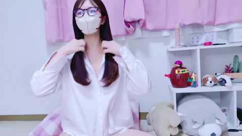 Media: Video of a young woman with straight brown hair, wearing glasses, white mask, and a white blouse, adjusting her hair in a pastel pink room with stuffed animals and shelves.