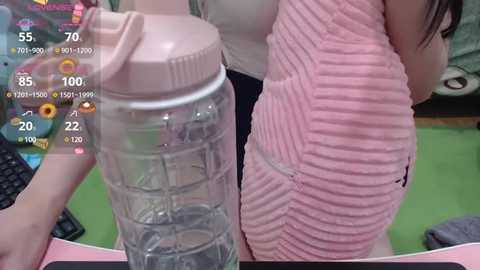 Video of two women, one wearing a pink ribbed sweater and the other a see-through water bottle, in a modern indoor setting with a green floor and keyboard.