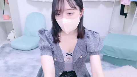 Media: Video of a young Asian woman with fair skin, short black hair, wearing a gray textured blazer over a white lace bralette, sitting on a soft, light gray rug. The room features a light blue chair and a white bed with a light blue sheet in the background.