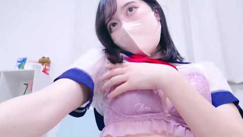 Media: Video of an Asian woman with pale skin, wearing a translucent, lace-trimmed pink bra and a sailor school uniform, with a white bandage over her mouth. Background includes a white curtain and a toy bear on a shelf.