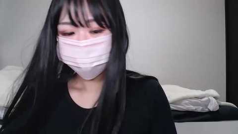 A video of a young Asian woman with long black hair, wearing a white face mask and black top, sitting on a bed with white and black sheets.