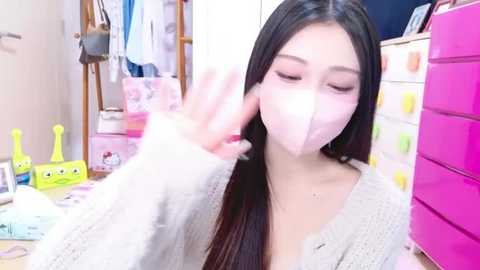 Media: Video of an Asian woman with long black hair, wearing a pink face mask, gray sweater, and a playful expression, waving. Background includes colorful storage boxes, a yellow SpongeBob SquarePants toy, and a pink dresser.