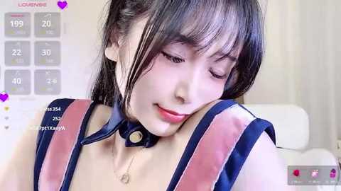 Media: Video of an Asian woman with dark hair and fair skin, wearing a blue choker and a blue and pink dress, sitting with her head resting on her hand, in a bright, indoor setting.