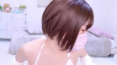 Media: Video of an Asian woman with straight, shoulder-length brown hair, wearing a white lace bra and a pink face mask, seated in a softly lit bedroom with white furniture and a plush teddy bear.