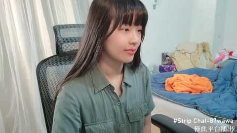 Media: Video of a young Asian woman with long black hair and bangs, wearing a green shirt, sitting in a black office chair, in a messy bedroom with a bed, orange blanket, and clothes.