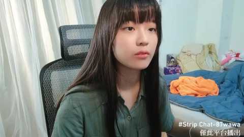 Media: Video of an Asian woman with long black hair, wearing a green shirt, sitting on a black office chair in a messy room with a bed and clothes scattered around.