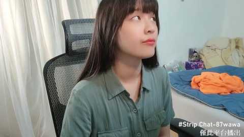 Media: A video of an Asian woman with straight black hair, wearing a green button-up shirt, sitting on a black mesh office chair in a cluttered bedroom with a bed, orange blanket, and white curtains.