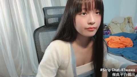 Media: Video of an Asian woman with long black hair, wearing a white shirt, sitting on a gray chair in a messy room with orange clothes on a bed.