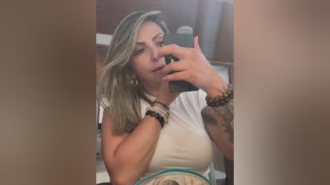 Media: Video of a woman with light skin and long, straight blonde hair, wearing a white tank top, taking a selfie with a smartphone in her right hand. She has multiple bracelets on her left wrist, and her expression is neutral.