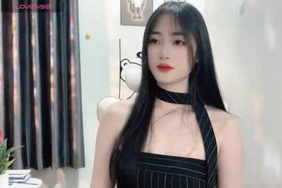 Media: Video of an East Asian woman with long black hair, wearing a black pinstripe halter dress, standing indoors against a white wall with curtains and a lamp.