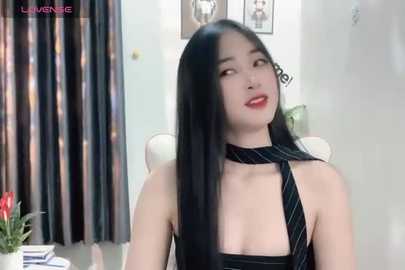 Media: Video of a young East Asian woman with long black hair, wearing a black halter top and striped tie, seated indoors with floral curtains, white walls, and framed photos in the background.