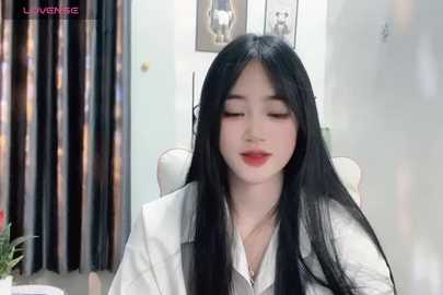 Media: A video of an Asian woman with long black hair, fair skin, and red lipstick, wearing a white blouse, seated in an office with framed art on the wall and curtains in the background.