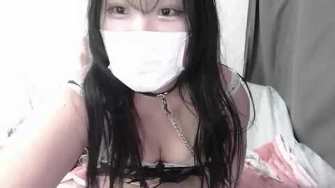 Media: Video of a pale-skinned woman with long black hair, wearing a white surgical mask, a black choker, and a revealing pink and white lingerie top. She is indoors with a gray curtain in the background.