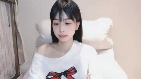 Media: Video of a young East Asian woman with straight black hair, wearing a white off-shoulder shirt with a red bow, sitting on a white cushioned chair against a plain beige wall.