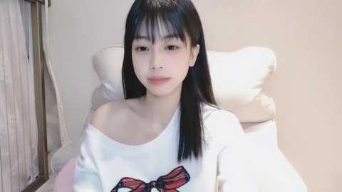 Media: Video of an East Asian woman with straight black hair and fair skin, wearing a white off-shoulder sweater with a butterfly graphic. She sits in a plush white chair against a beige wall.