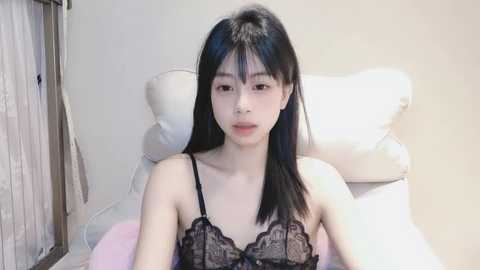 Media: Video of an East Asian woman with straight black hair, wearing a black lace bra, sitting on a white cushioned chair in a minimalist room with beige walls.