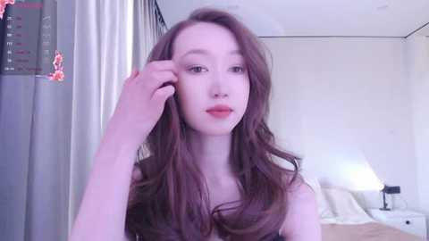 Video of a young Asian woman with fair skin, long brown hair, and a delicate face, seated in a modern bedroom with white walls, a bed, and a lamp.